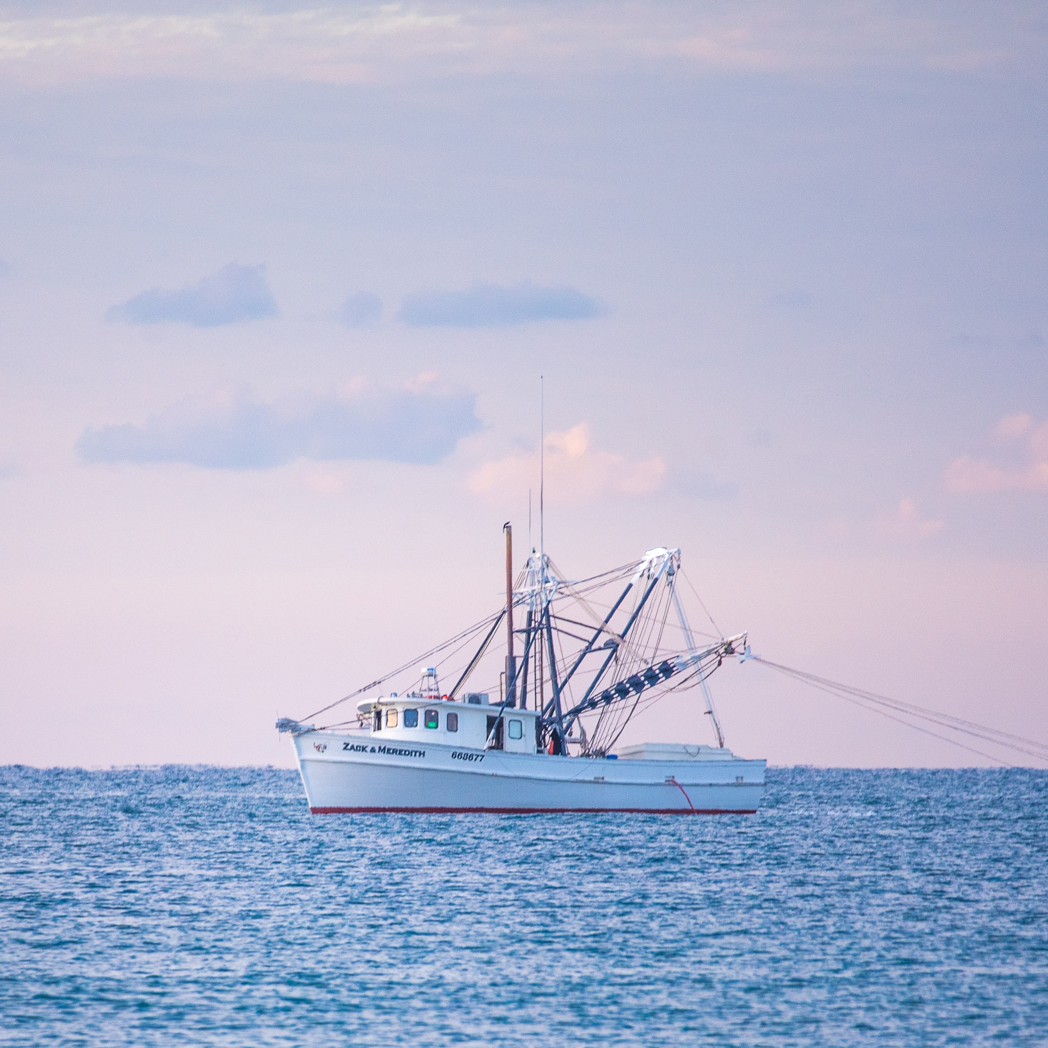 Crevettier blanc, le ZACK & MEREDITH, sur mer agitée au crépuscule. Le ciel est rose et lavande. Outil de pêche visible.