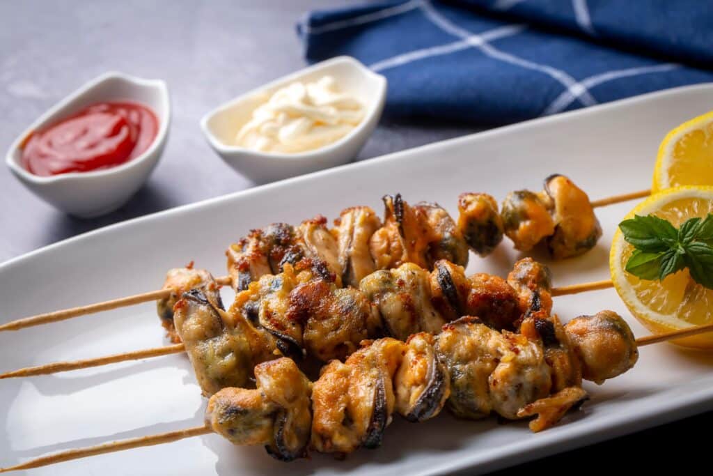 Brochettes de moules grillées : le parfait apéritif marin. Brochettes de moules grillées dorées, servies sur plat blanc avec citron, menthe, et deux ramequins de sauces (ketchup et mayonnaise).