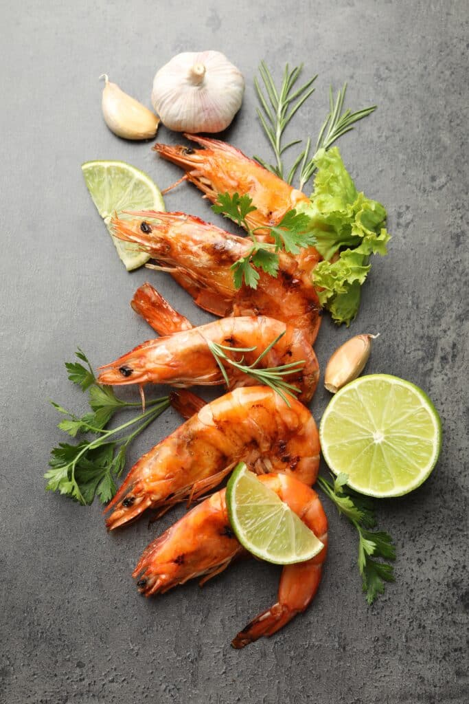 Crevettes grillées à l'ail et au citron vert pour un repas savoureux. Cinq grandes crevettes grillées, garnies d'ail, de tranches de citron vert, de persil et de romarin, sur fond de pierre grise.