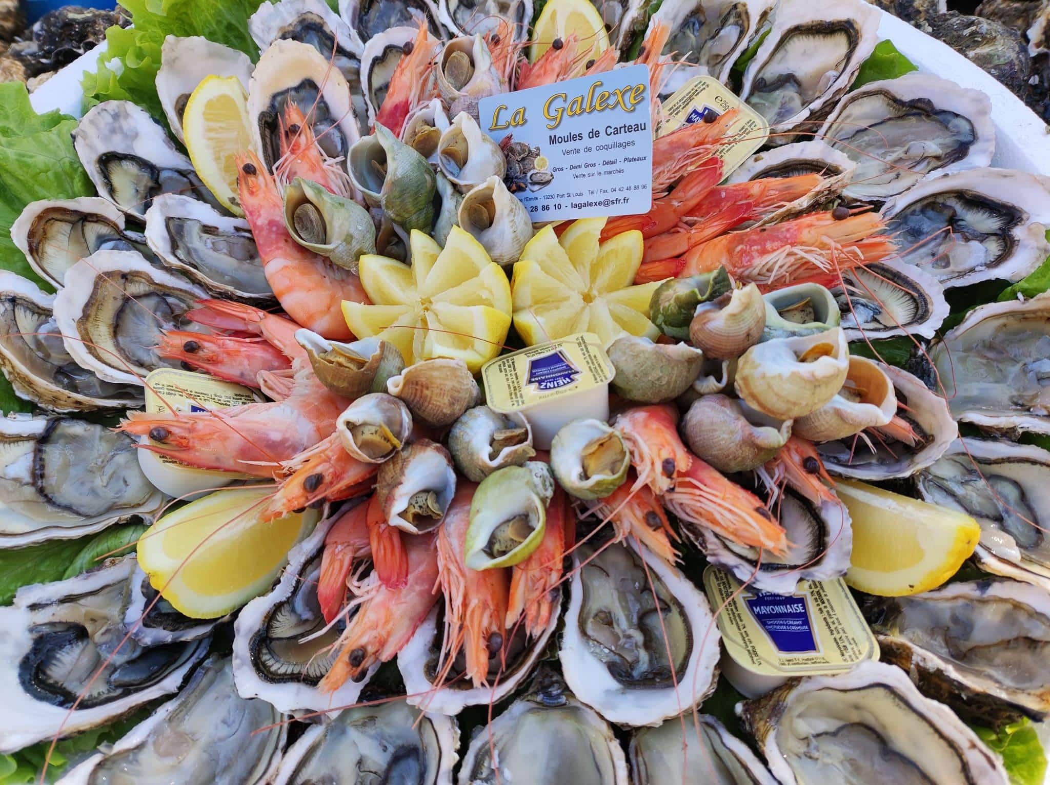 Plateau abondant de fruits de mer frais: huîtres, crevettes, bulots, citron et mayonnaise. Carte La Galexe Moules de Carteau visible.